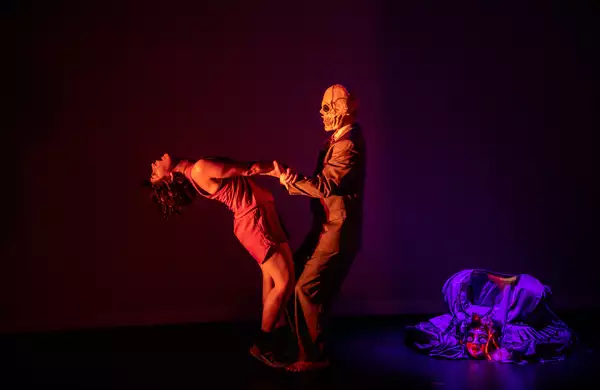 Two people dance with a puppet in the background. Oner person wears a skull mask while the other has their head thrown back.