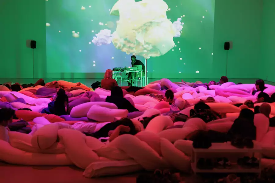 A listening session showing a group of people lying on large, colorful, soft cushions in a dimly lit room, watching a digital visual display projected on the wall. The lighting transitions between soft pinks and greens. At the front, Ashley Holmes sits at a desk with electronic equipment.