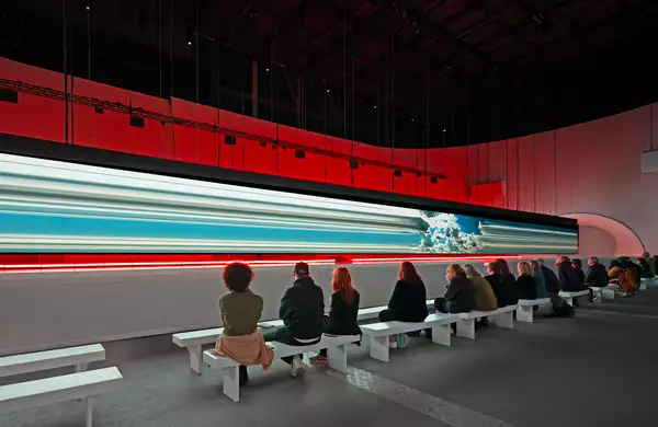 Spectators sit on a bench watching a long horizontal screen suspended over a white catwalk