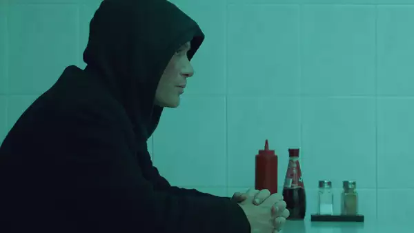 The image shows a person sitting at a table in what appears to be a dimly lit, minimalist diner or café. The person is wearing a dark hoodie with the hood up, facing left, and sitting with their hands clasped in front of them, creating a contemplative or introspective mood. On the table in front of them, there are bottles of ketchup, vinegar, and salt and pepper shakers, adding to the simplicity of the setting. The greenish-blue tint of the lighting adds a moody, cinematic atmosphere to the scene.