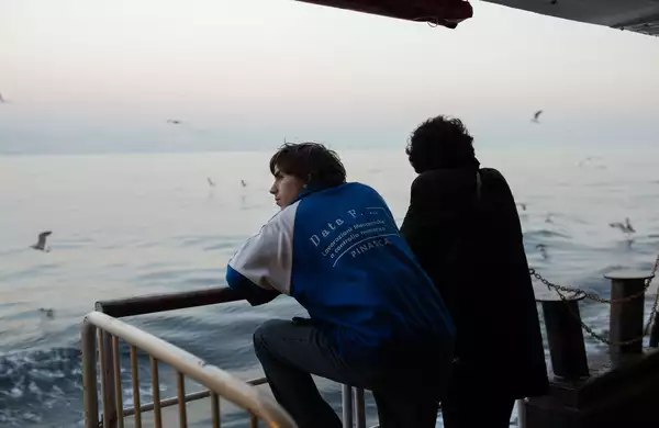 Still from Crossing. Two characters with their backs turned, stood by railings looking out at sea.