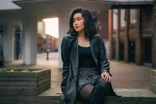 A woman is sitting on a wooden bench, body facing towards the camera but she is looking off to the left into the distance. She is wearing a long black jacket, black top and flowery skirt.