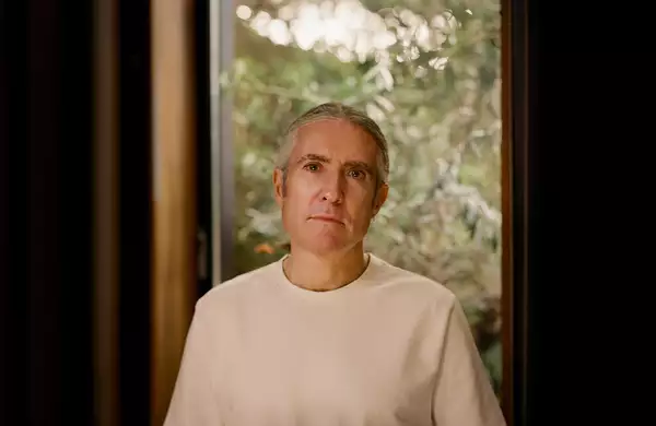 A portrait of Richard Russell wearing a light-coloured shirt, standing in front of a window with a blurred green outdoor background.