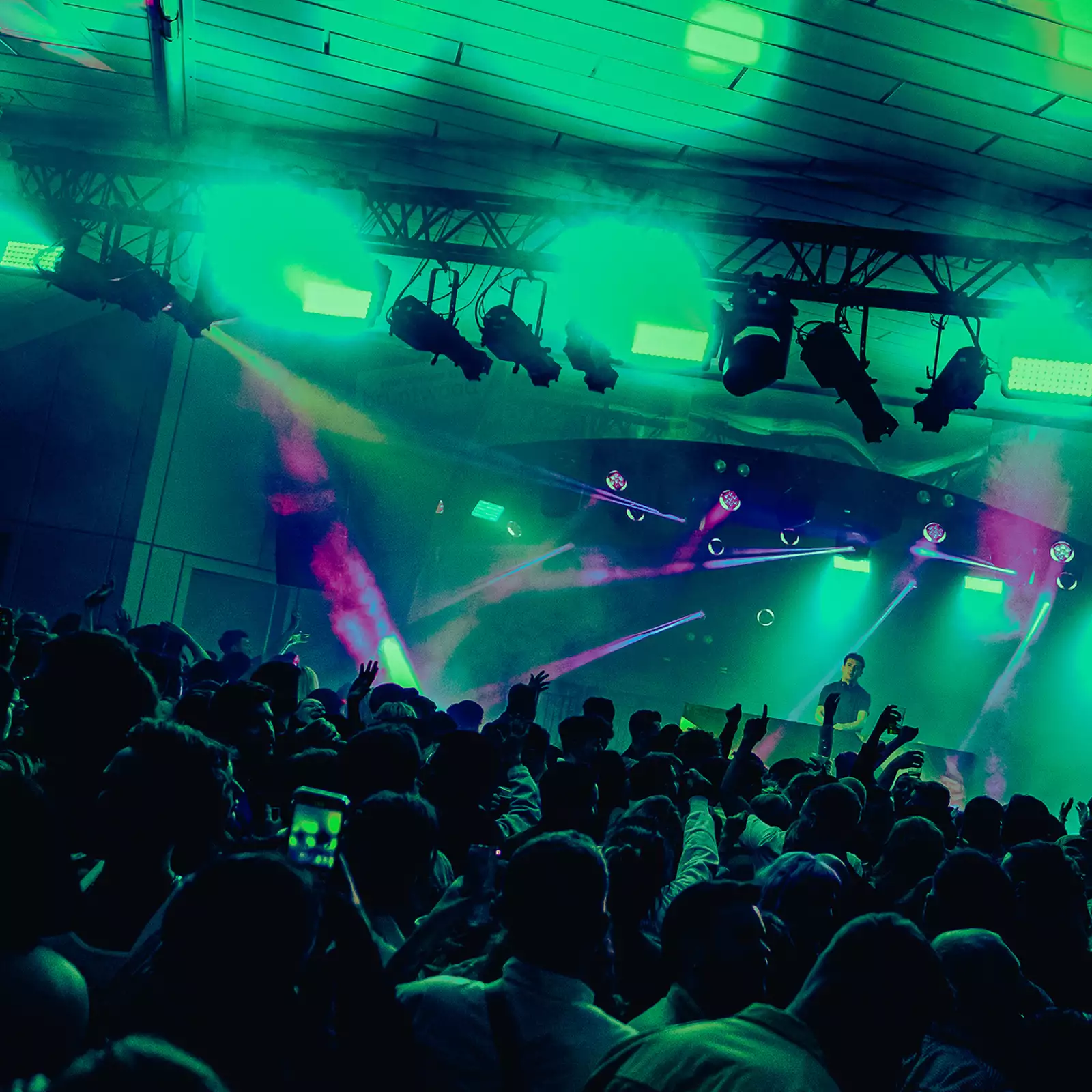 A DJ set on Festival Square from MIF23, showing a large crowd of people dancing and filming a DJ on stage. The lighting is hazy green and there are purple strobe lights.