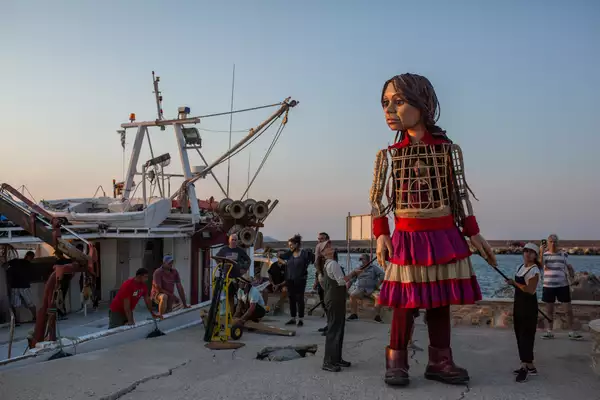 The Little Amal puppet supported by two puppeteers in overalls and grey caps as well as a puppeteer inside. She looks over curiously at a fishing boat. A handful of people are stood on and around the boat, with a few people sat on a short stone wall next to the sea.