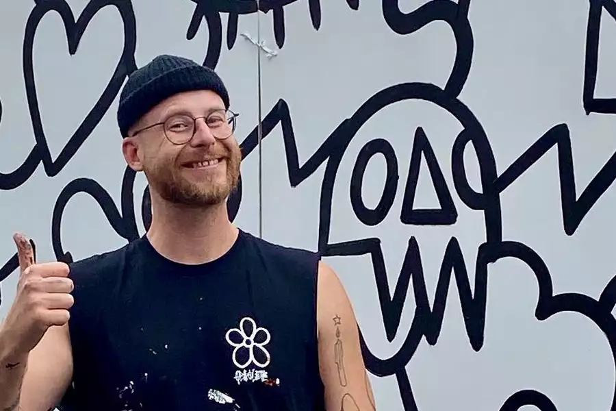 Pete Obsolete smiling with their thumbs up, wearing a black beanie and stood in front of a doodle wall