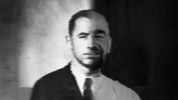 Black and white photo headshot of a person. One half of their body is clean-shaven and wearing a suit and tie; the other half is not.