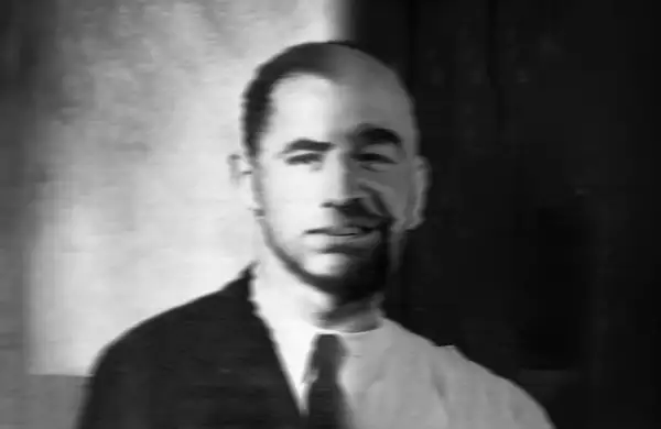 Black and white photo headshot of a person. One half of their body is clean-shaven and wearing a suit and tie; the other half is not.