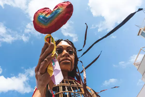 Close-up of the Little Amal puppet holding up a giant rainbow lollipop in the shape of a heart, made out of papier-mâché