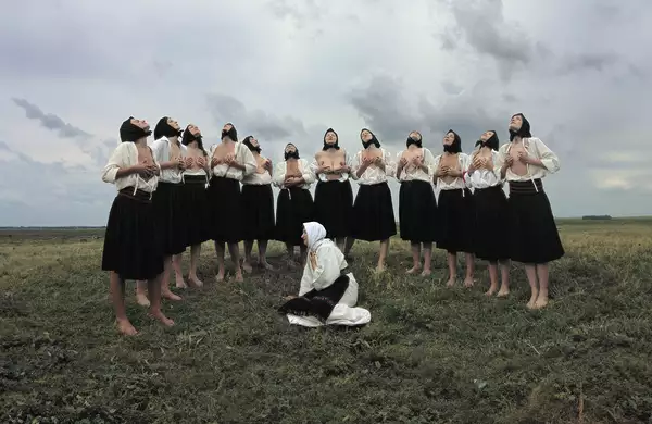 A group of women in traditional Eastern European attire stand barefoot in a grassy field under a dramatic, cloudy sky. Most are wearing black skirts, white blouses, and black headscarves, with some partially exposing their chests while lifting their heads upward in a ritualistic pose. In the foreground, a single woman dressed in white kneels on the ground with a headscarf and embroidered belt, gazing to the side.