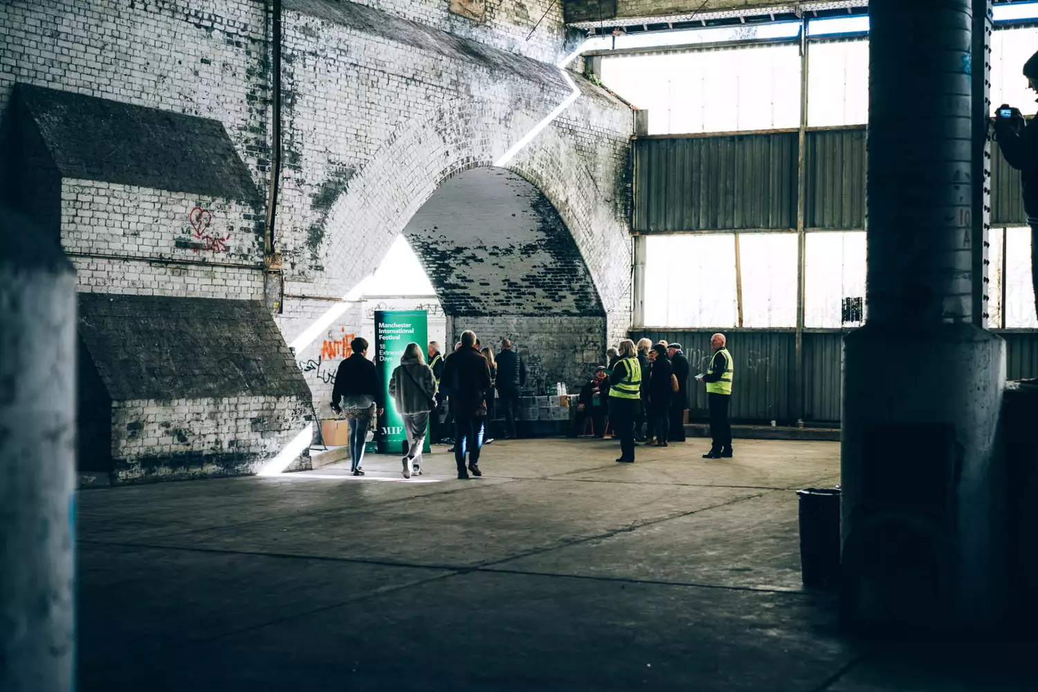 A small crowd of people are gathered in a large room with a brick facade on one side and windows on the other.