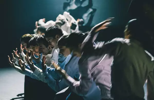 A group of dancers stood in a line with their arms extended forward, moving in blurred motion. The lighting is dim, casting dramatic shadows.