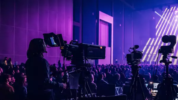 A camera operator films a live event in the Warehouse, which is filled with people. There is dramatic purple lighting with multiple cameras and bright stage beams.