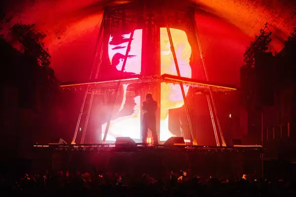 A photograph of Skepta, stood in silhouette on a brightly lit stage. The stage is a custom-built pyramid with a flat top, with a centrepiece screen showing 3D graphics of a human head shown across multiple large LED displays