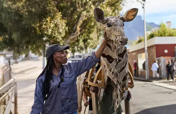 A woman in a cap touches the head of a life-sized giraffe puppet made from cardboard and wood, as part of The Herds. The background shows an urban environment with people watching and a scenic view of trees and mountains.