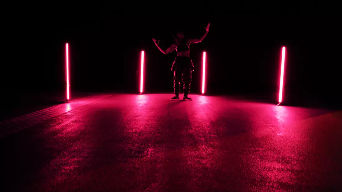 A person stands in a dimly lit space between four vertical neon lights emitting a pink glow, their arms raised in a dramatic pose