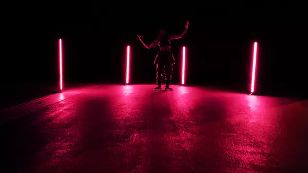 A person stands in a dimly lit space between four vertical neon lights emitting a pink glow, their arms raised in a dramatic pose