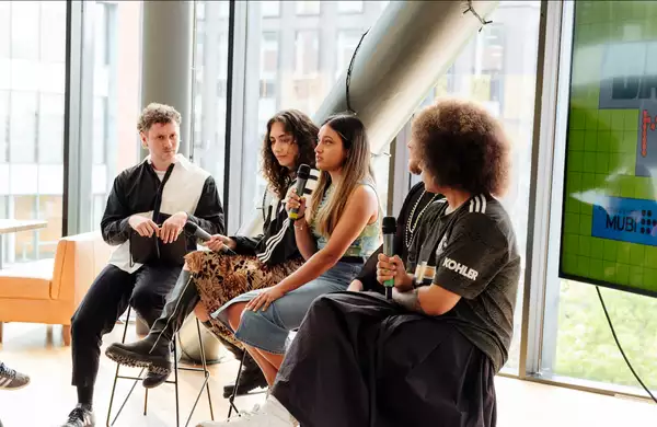 Four people sat on high chairs in front of a large window during a panel talk. One is speaking. There is a screen behind them with Dazed and Mubi branding on.