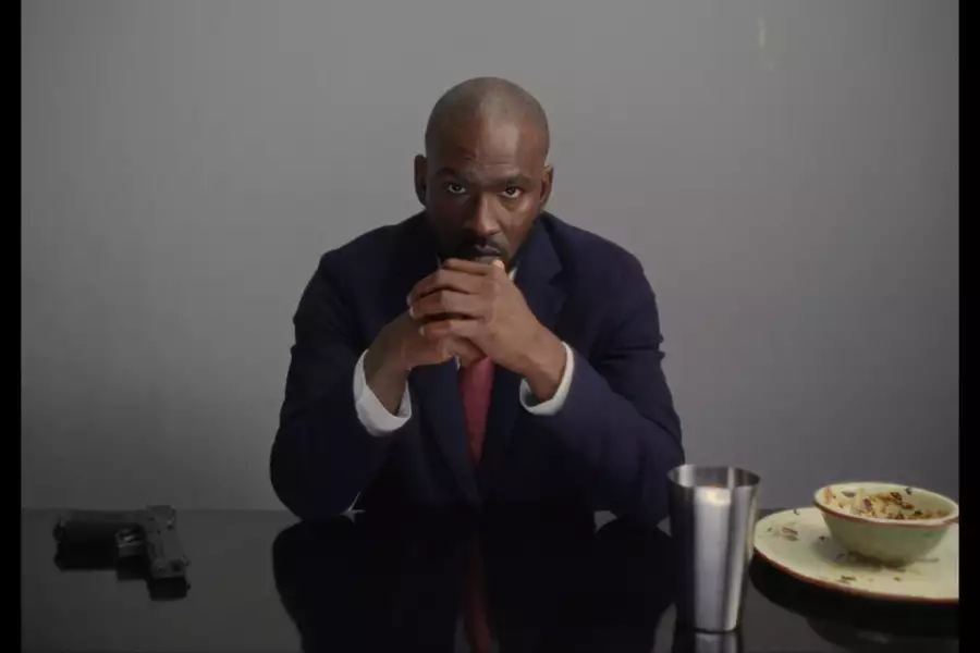 Still from Tribal Mark showing Skepta sitting at a table with dirty dishes and a gun on it