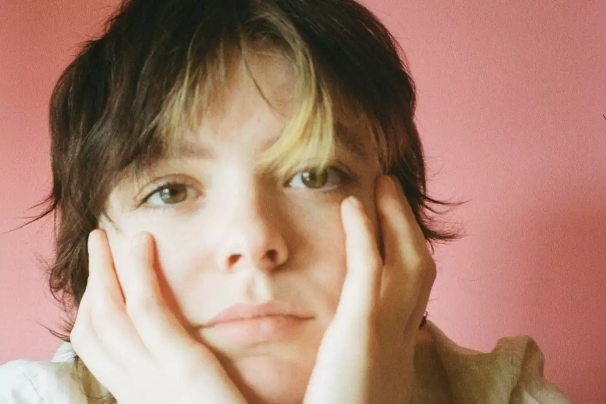 Close-up headshot of Olivesque, with their head resting on their hands. The background is pink.