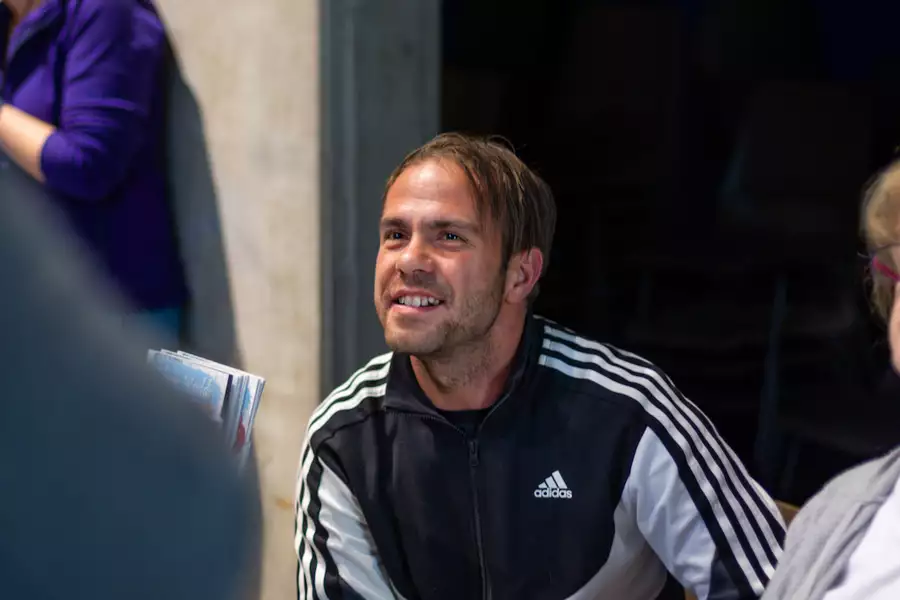 A smiling man wearing a black and white Adidas track jacket is seated indoors, engaging with someone off-camera.
