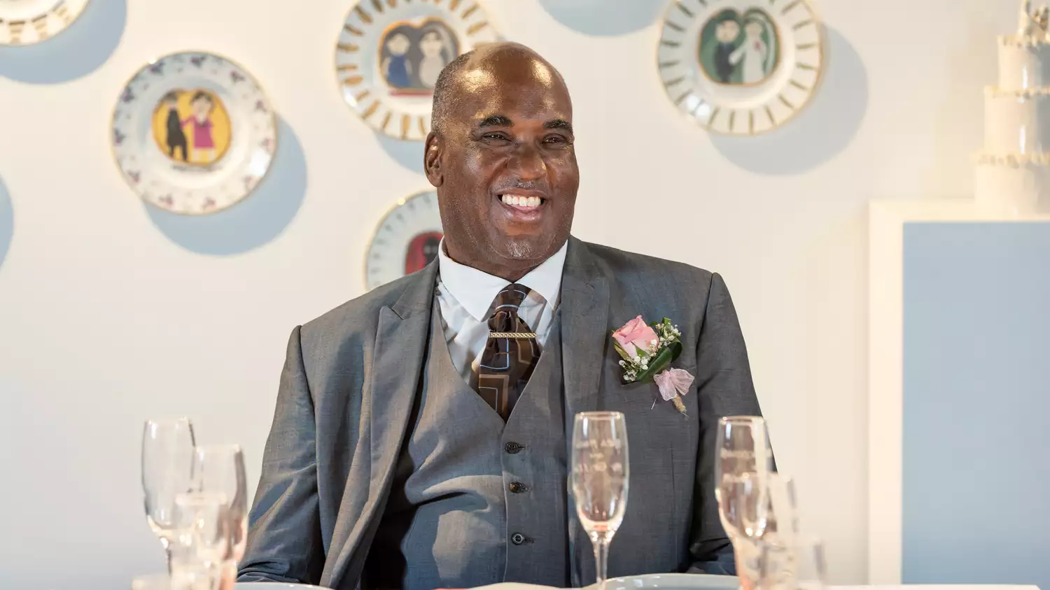 Horace Lindezey in a suit with a pink rose sits at a table with champagne glasses, in front of a wall decorated with illustrated plates.