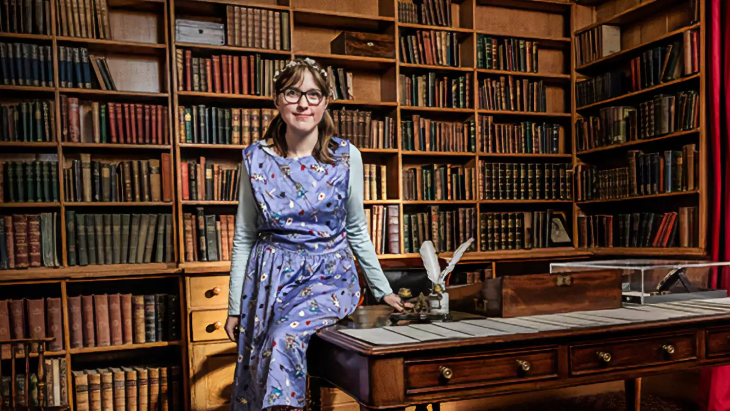 Sally Hirst wearing a blue patterned dress, smiling and perched on an old wood desk in a library.