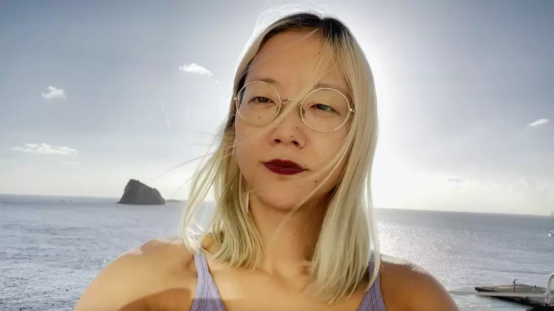 A photograph of Christine Sun Kim, backlit by the sun and standing in front of the sea with a small island above her right shoulder