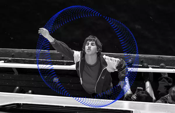 A still from the film Crossing. A male character wearing a tracksuit jacket is leaning off the edge of a boat and reaching up to the sky with his eyes closed.