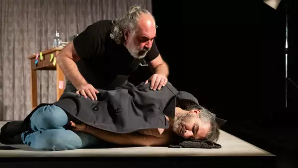 A person lying down on stage with eyes closed underneath a coat – with another person peering over him